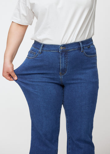 Dark blue mid-rise bootcut jeans in Oceans. A plus-size model stretches the jeans at the thigh to show the fabric&