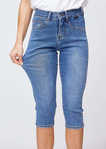 Slim capris for women in blue denim, color Rivers. A model pulls the fabric at the thigh to demonstrate stretch and flexibility.