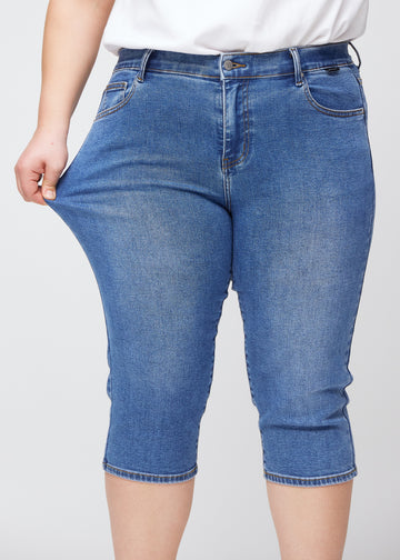 Slim capris for women in blue denim, color Rivers. A plus-size model pulls the fabric at the thigh to demonstrate stretch and flexibility.
