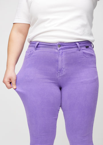 Skinny jeans for women in purple, color Lavenders. A plus-size model stretches the jeans at the thigh to show the fabric&