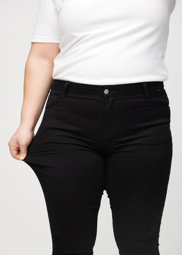 Skinny jeans for women in black, color Ravens. A plus-size model stretches the jeans at the thigh to show the fabric&