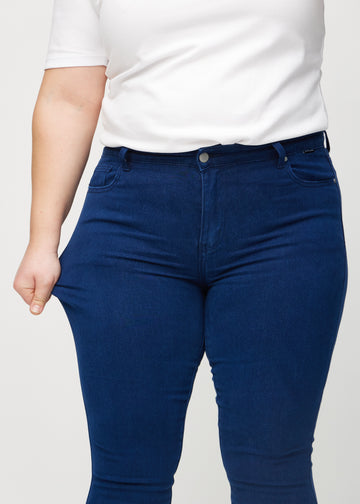 Skinny jeans for women in dark blue, color Royals. A plus-size model stretches the jeans at the thigh to show the fabric&