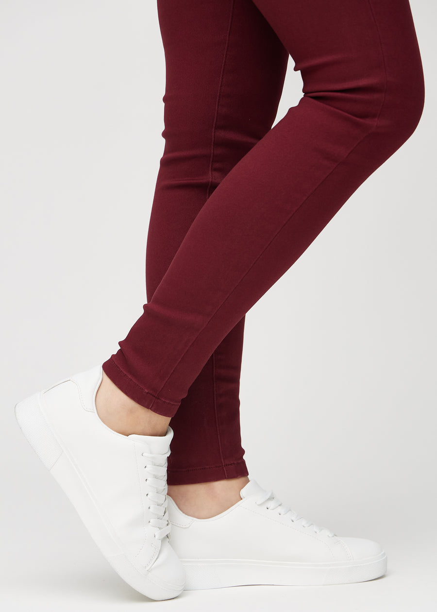 Skinny jeans for women in dark red, color Santas. Close-up of the snug-fitting leg openings at the ankles on a model.