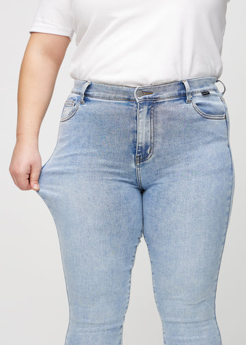 Skinny jeans for women in light blue, color Waves. A plus-size model stretches the jeans at the thigh to show the fabric&