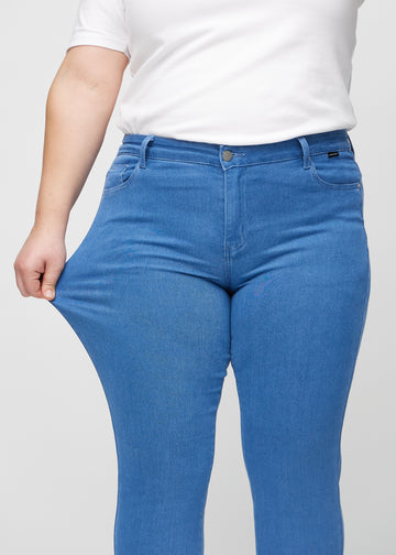 Slim jeans for women in blue, color Geraniums. A plus-size model stretches the jeans at the thigh to show the fabric&
