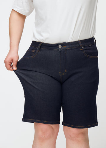 Plus-size model stretches deep dark blue denim shorts for women in the colour Midnights at the thigh to show stretch.